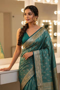 Woman in a teal saree with gold patterns standing in front of a mirror.