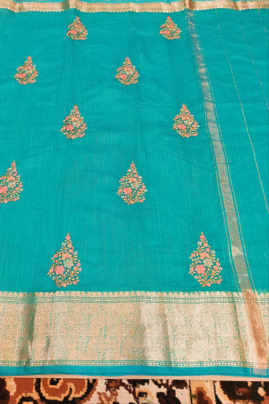 Turquoise saree with gold and pink floral patterns on a patterned fabric background