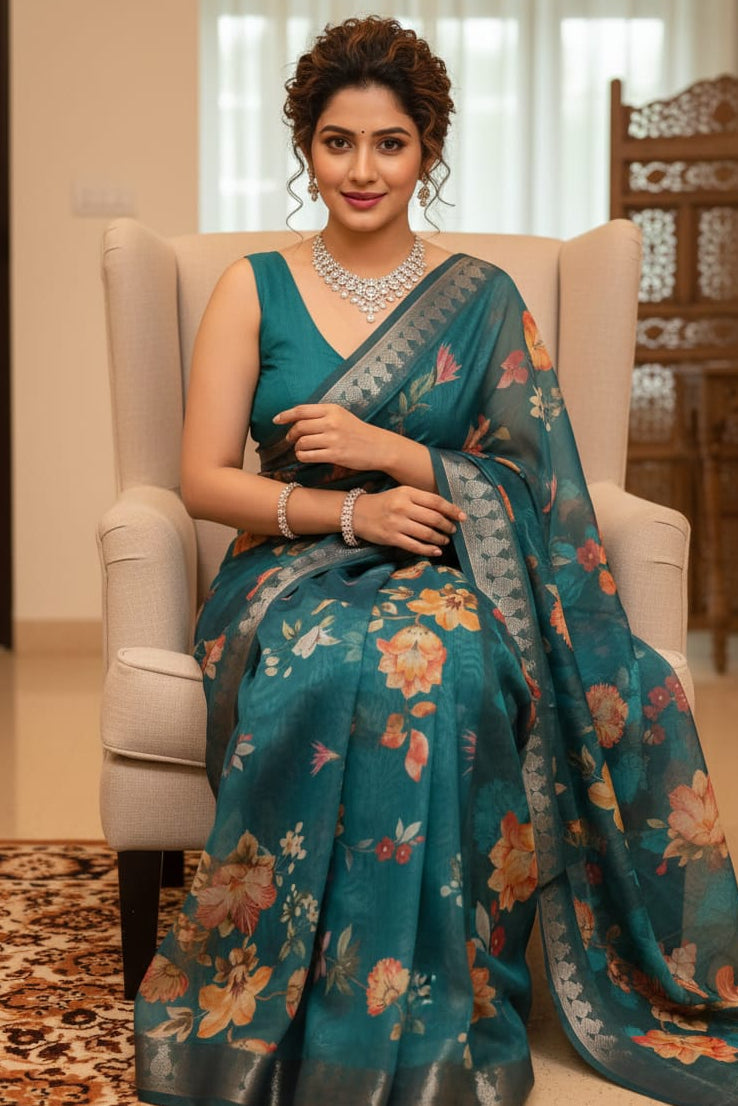 Woman in a teal saree with floral patterns sitting on a chair indoors.