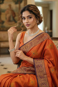 Woman in an orange saree with intricate patterns in a decorated room.
